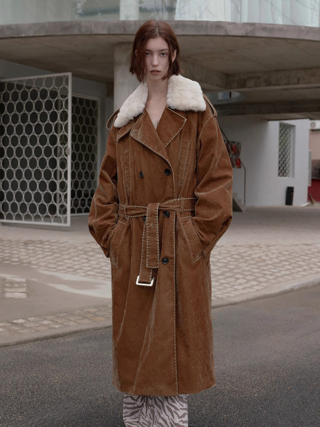 Person wearing a brown coat with white fur collar standing on a street.